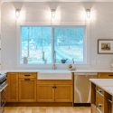 Kitchen sink wall with farmhouse sink, Lectus Beckham warm maple shaker base cabinets, stainless steel dishwasher, white quartz countertops, subway tile backsplash, and bright window lighting in a Nanaimo, BC kitchen renovation